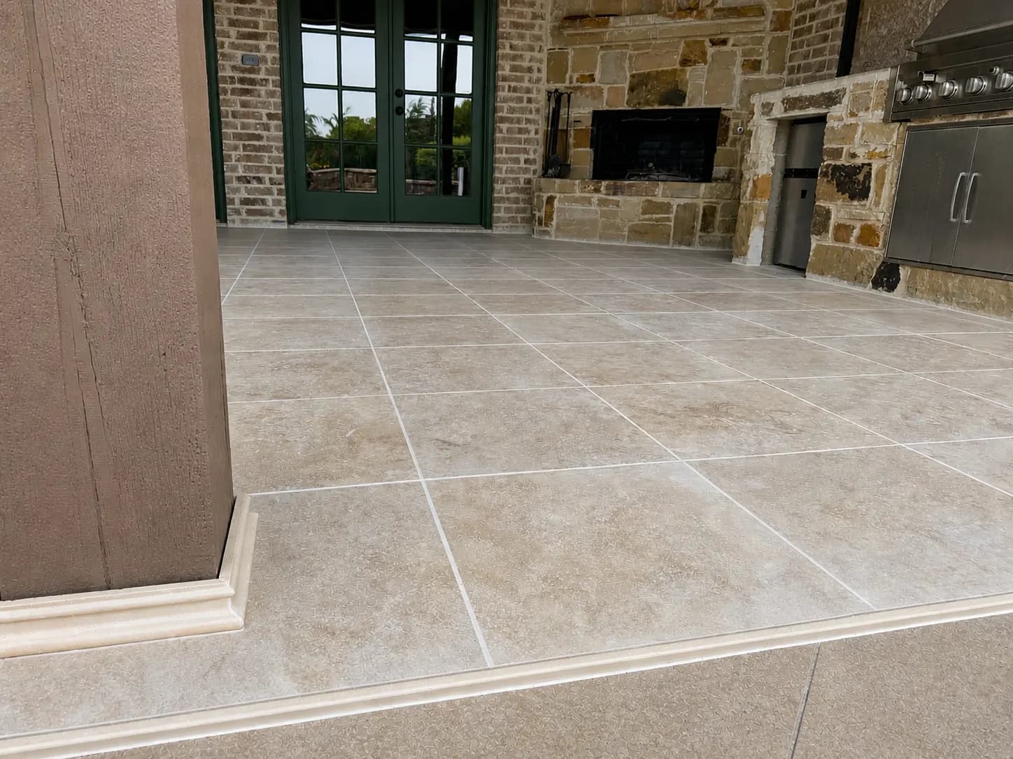 Travertine tile patio with stone-clad outdoor fireplace and built-in grill, French doors leading inside.