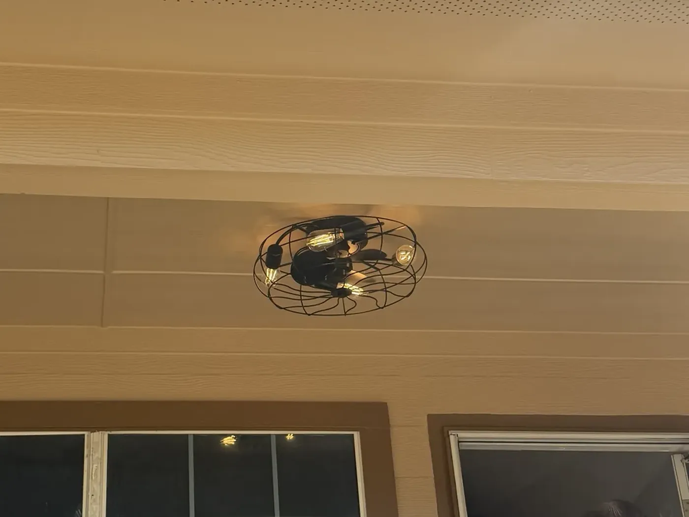 Black caged ceiling fan with Edison bulbs mounted on a tan porch ceiling, lit after dark.