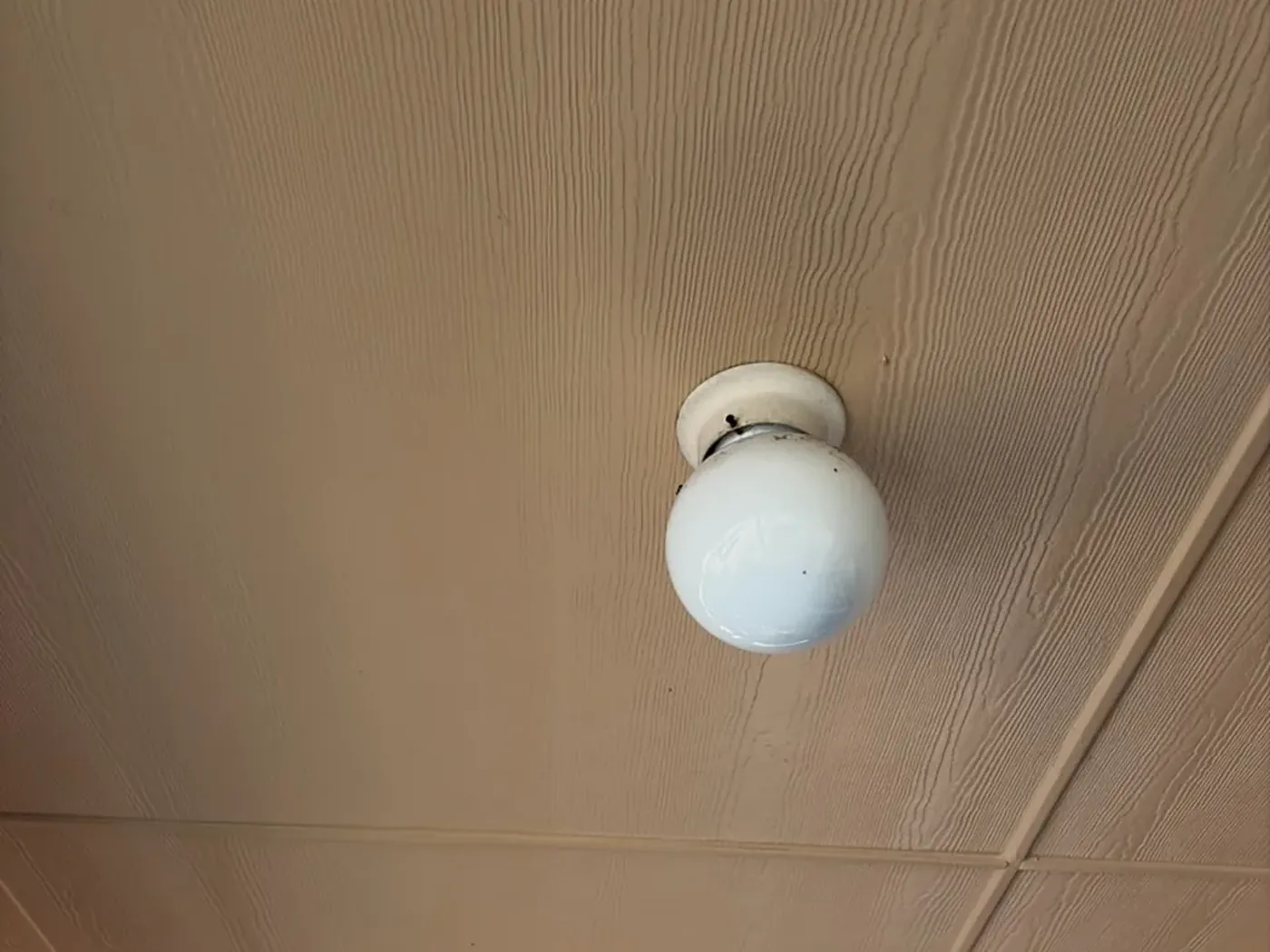 A dated white glass globe light flush-mounted to a tan panelled porch ceiling.